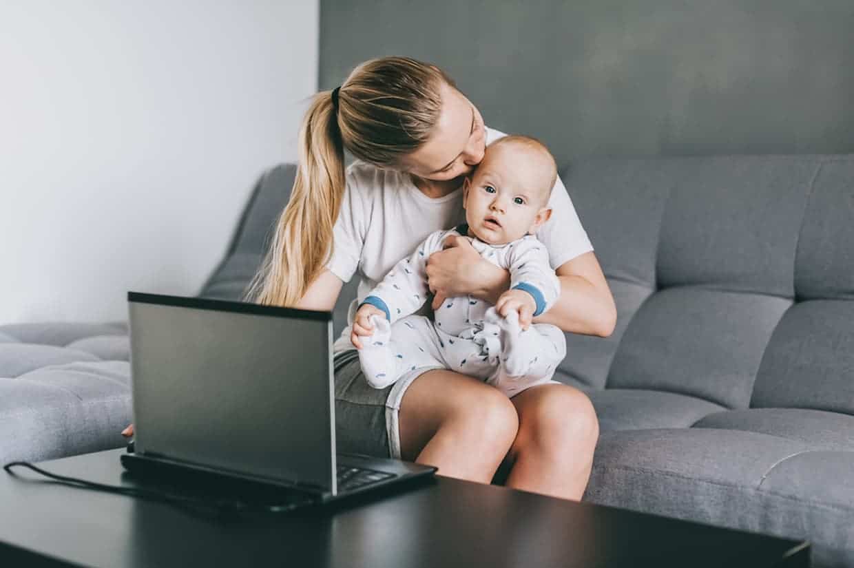 Young mom kissing her infant child while working at her laptop at home.