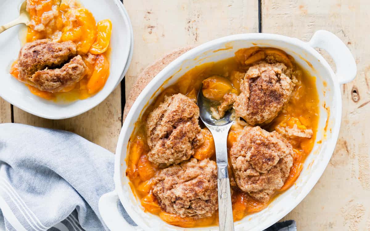 Apricot cobbler in a white baking dish with wooden metal serving spoon on a wooden surface.
