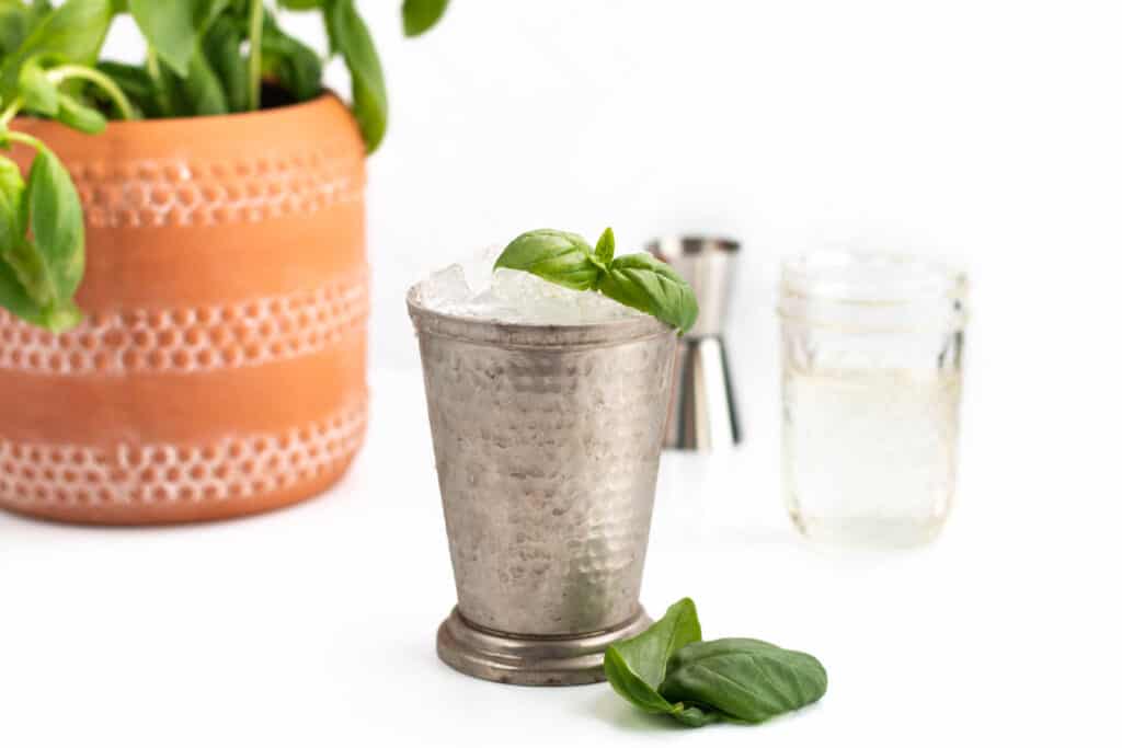 A metal julep cup stands in the center of the photo with a green leaf garnish. Behind it sits a terra cotta pot with basil growing, a mason jar of simple syrup and a metal cocktail jigger