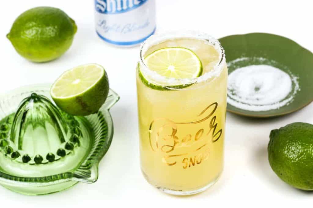 A beerita with a salt rim in a glass shaped like a beer can next to limes and a green glass juicer.