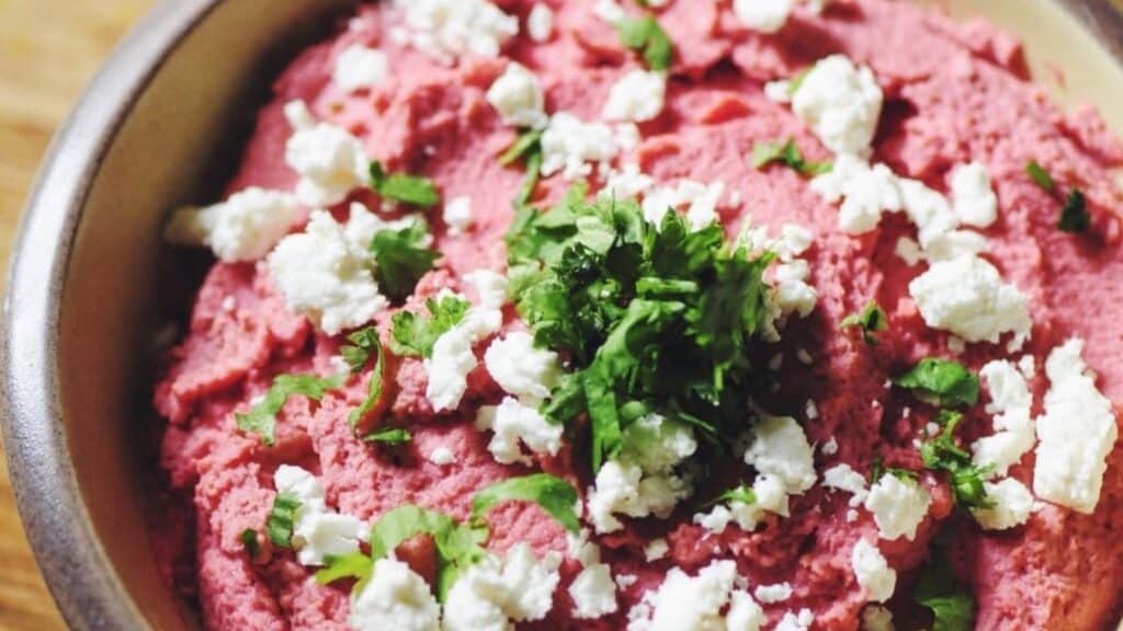 A bowl filled with creamy pink hummus topped with feta cheese and green herbs.