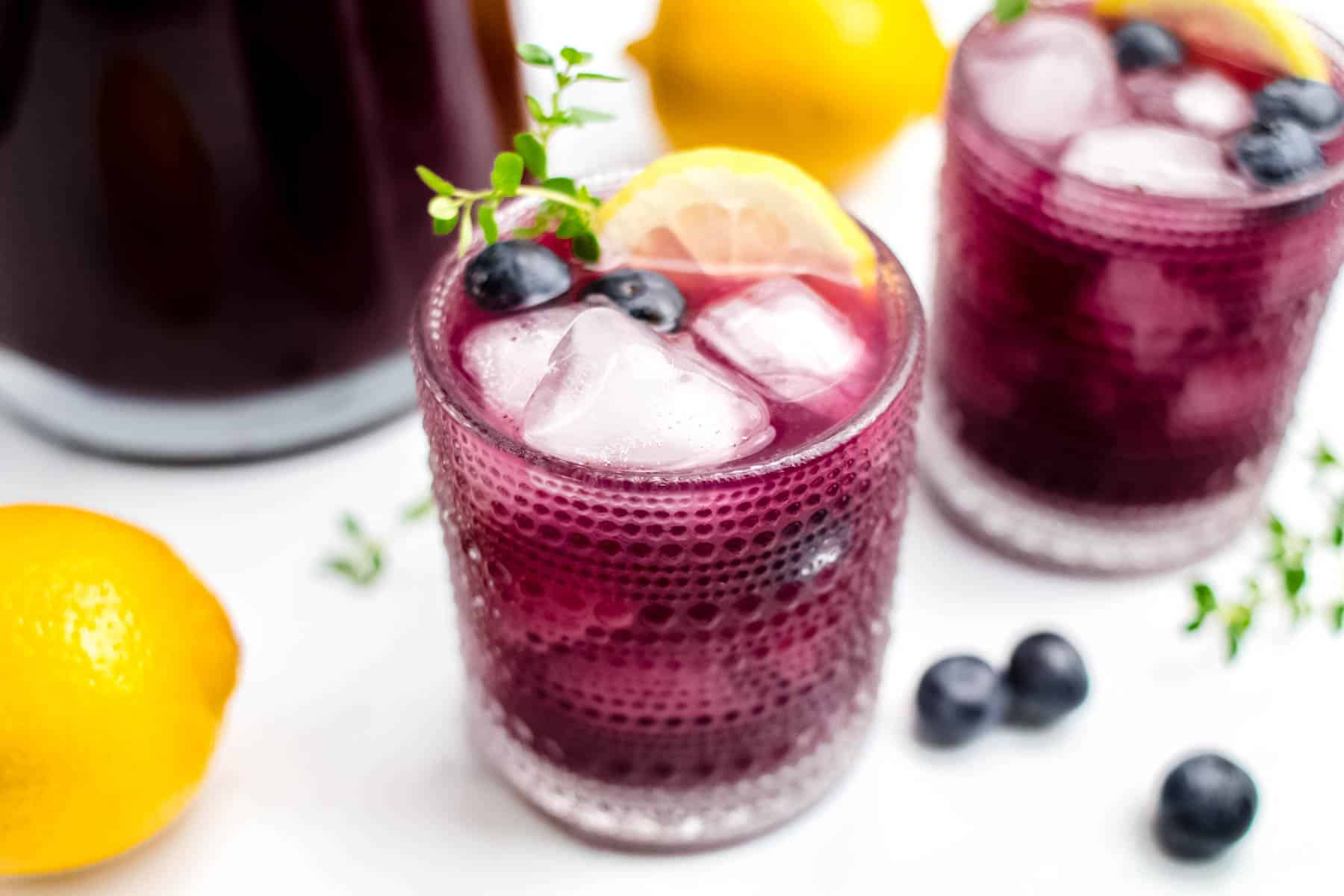 Two glasses of blueberry lemonade garnished with lemon, blueberries and herbs.