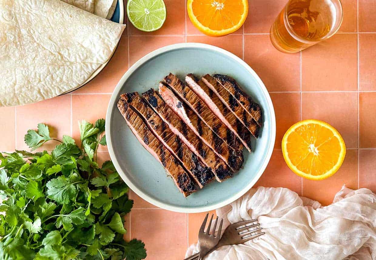 Carne asada on a light blue plate surrounded by oranges, limes, tortillas, cilantro and beer.