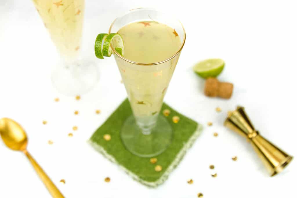 A champagne version of a moscow mule in a glass rests on a green coaster surrounded by gold cocktail tools and half a lime.