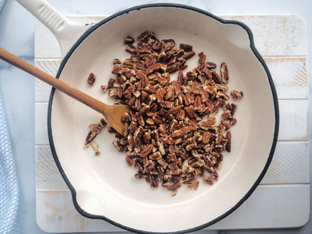 A white skillet on a white board with chopped pecans and a wooden spoon.