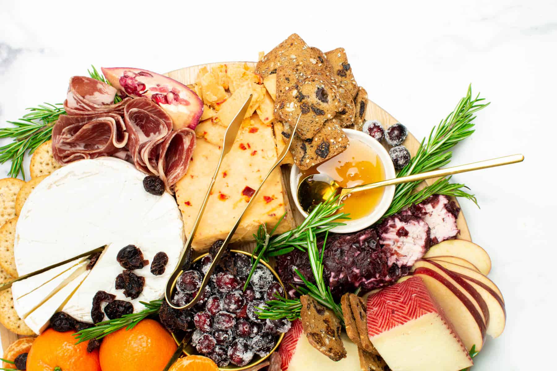 Cheeses and meats are arranged decoratively on a round wooden plate with spreads and fruits among them.