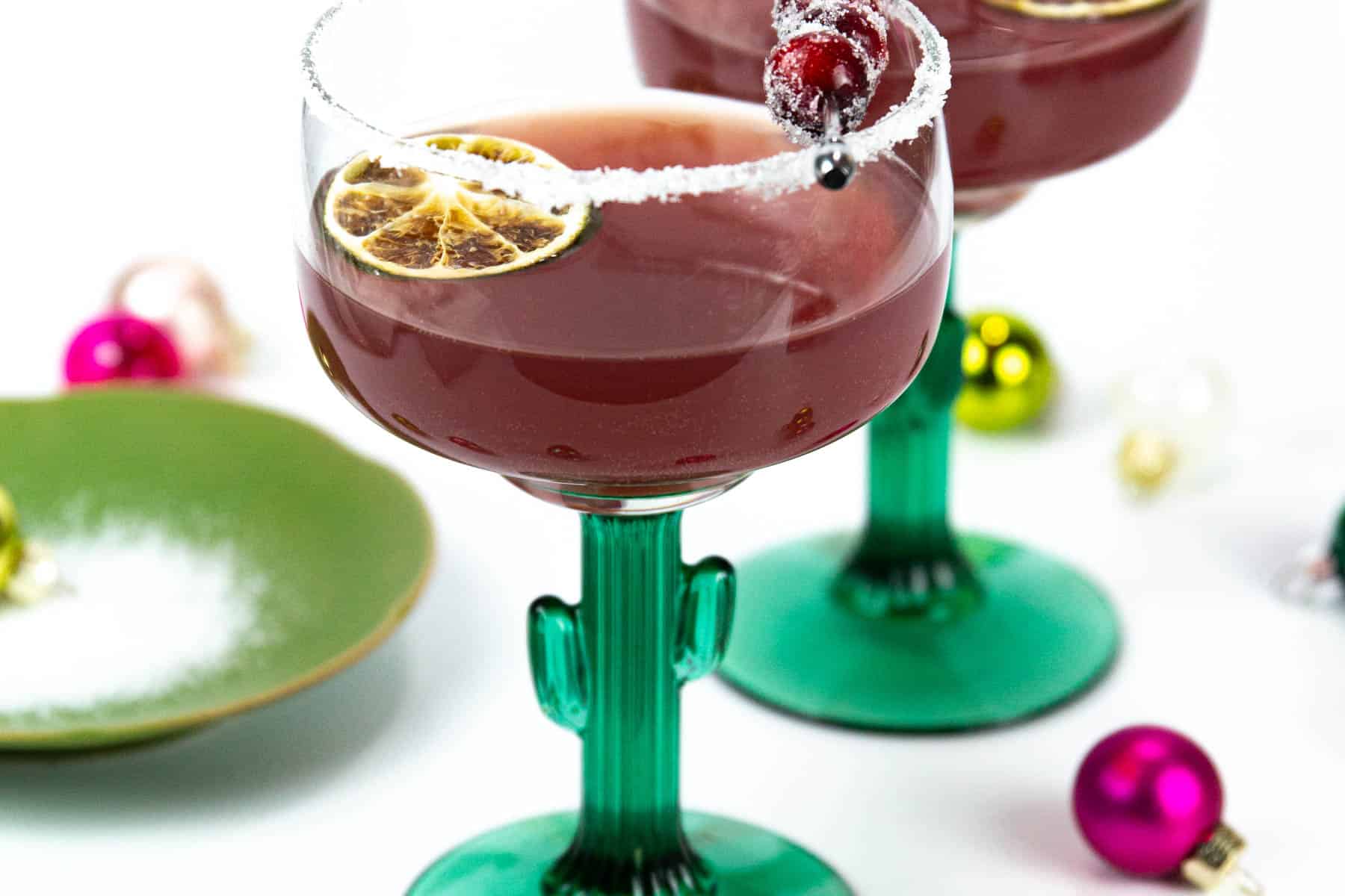 Two cranberry margaritas next to Christmas ornaments and a plate of salt.