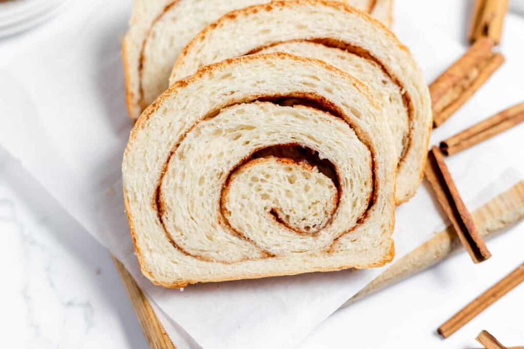A slice of cinnamon swirl bread with other slices behind it and cinnamon sticks to the side. 