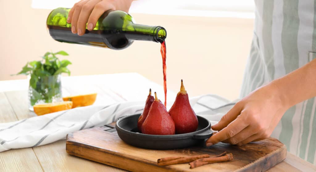 Woman pouring wine onto pan with sweet pears in kitchen.