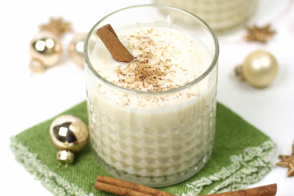 A glass of Dairy-Free Eggnog with a cinnamon stick and a sprinkle of grated nutmeg on a green linen coaster.