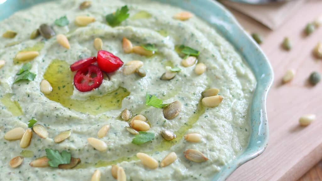 Roasted eggplant dip drizzled with olive oil and topped with pine nuts and parsley in a teal dish.