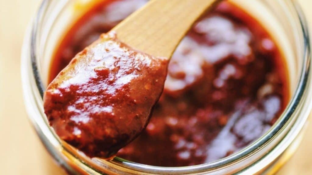 A close shot of a wooden spoon scooping red sauce out of a glass jar.