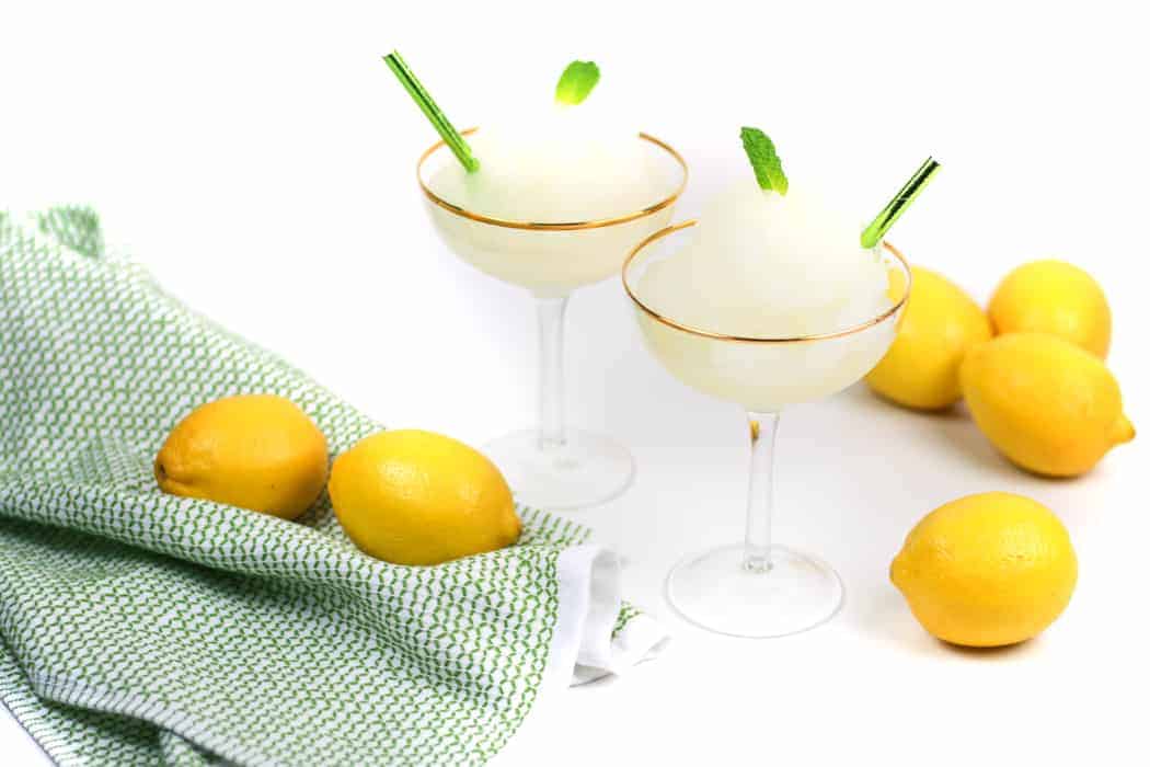 Lemon Daiquiris in coupe glasses garnished with mint leaves and surrounded by lemons and a green kitchen towel.
