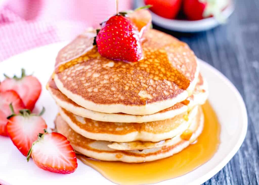 A stack of gluten free pancakes on a plate garnished with strawberries and being drizzled with maple syrup.