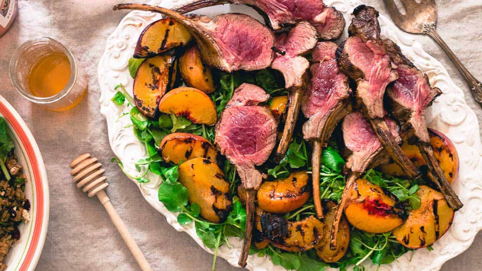 Grilled lamb chops with peaches over baby greens on a white serving platter.