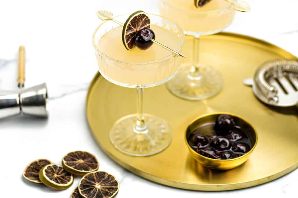 A gold tray holds two rum cocktails in coupe glasses, the lid to a cocktail shaker and a bowl of cherries. additional garnishes and tools sit beside on a white marble surface.