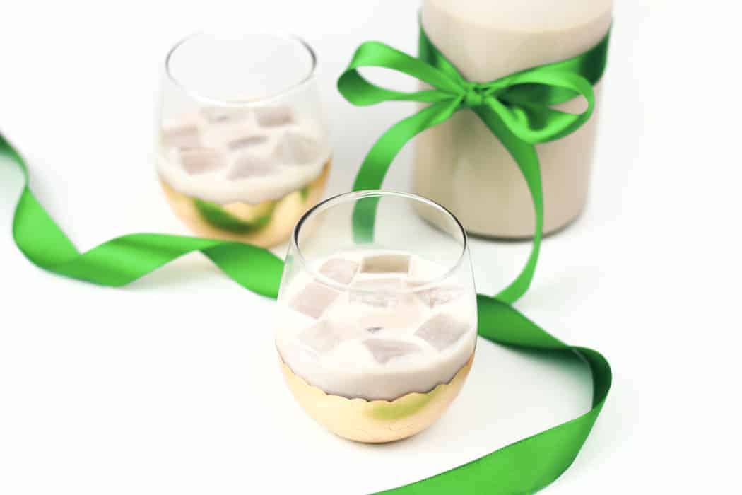 Two glasses of Irish cream on the rocks next to a bottle tied with a green ribbon for gifting.