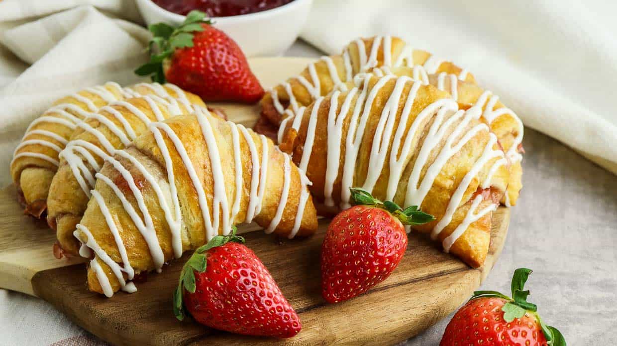 Jam crescent rolls drizzled with white icing with strawberries scattered around.