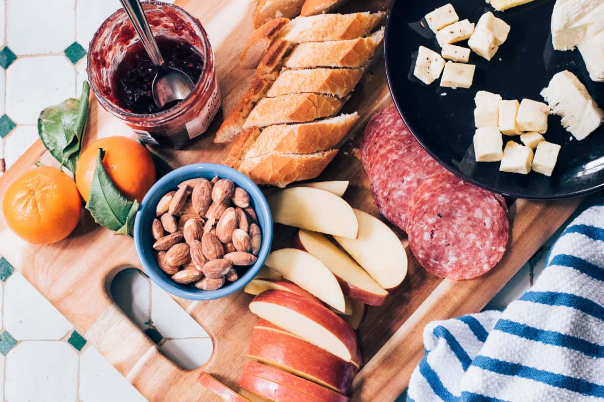 An orange, a jar of jam, meats, apples, nuts and cheeses on a grazing board.