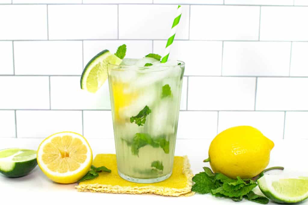 Lemons and limes sit alongside a glass of lemon-lime mojito with mint leaves and a green and white striped straw