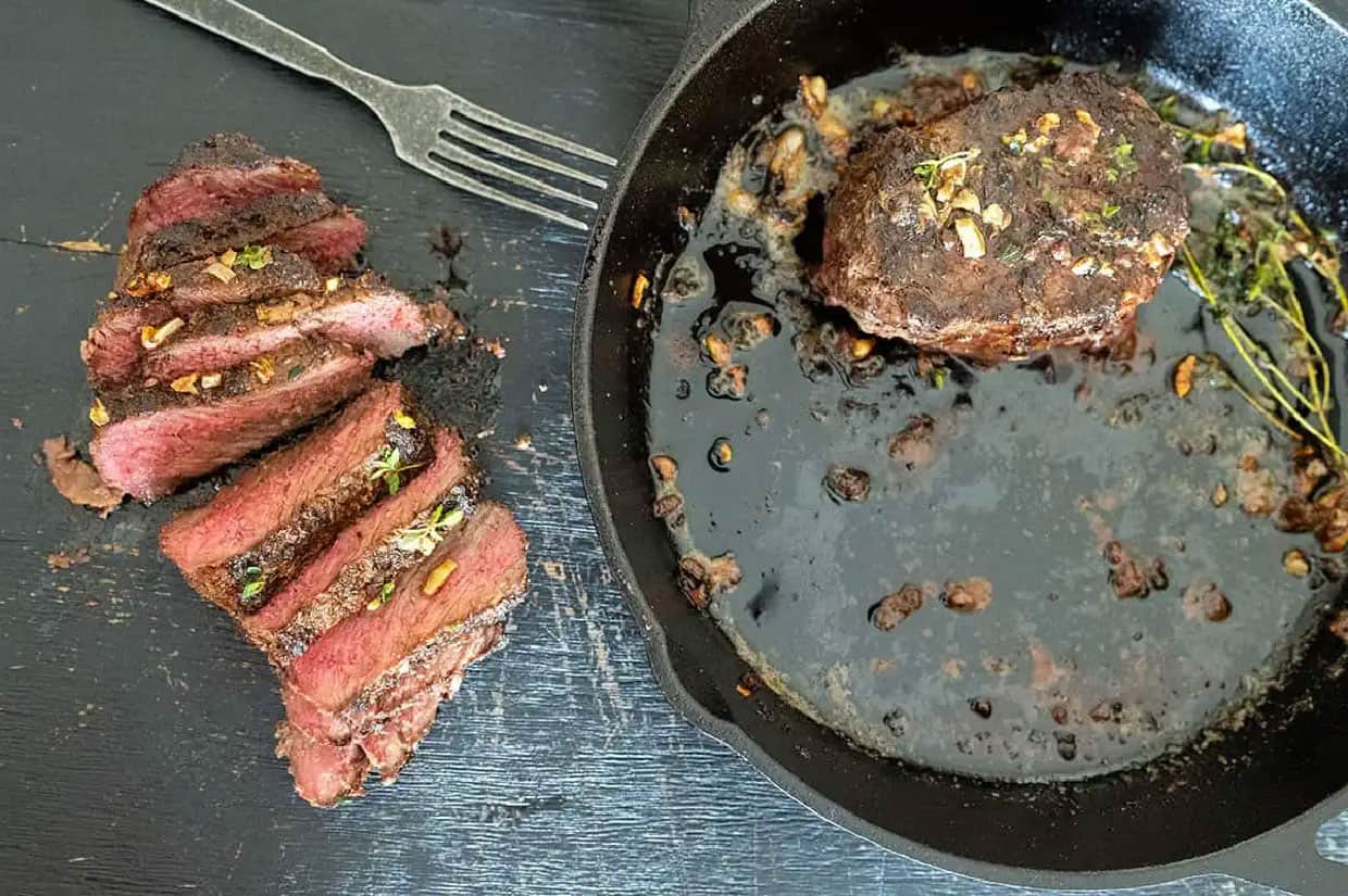 sliced medium-rare steak next to cooked steak in cast iron skillet.