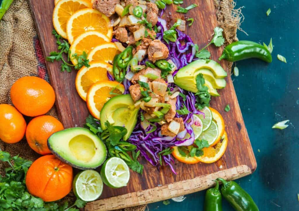 Pork bites on a cutting board with colorful fruits.