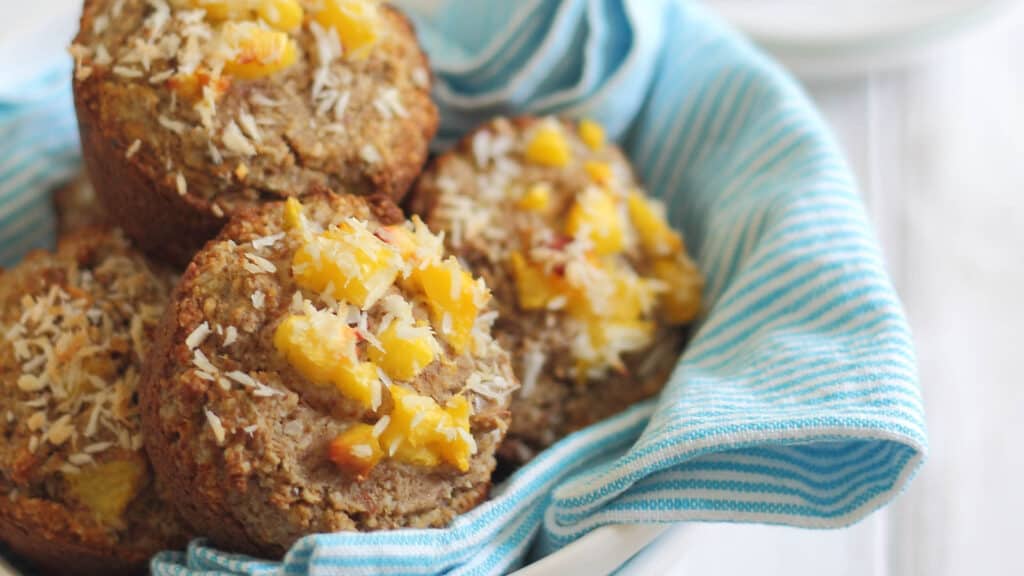 Almond flour peach coconut muffins in a bowl lined with a blue striped kitchen towel.