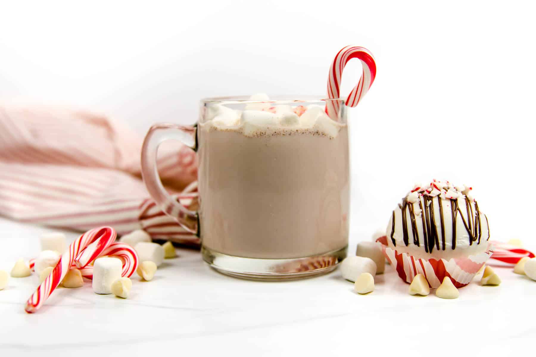 Ingredients for a peppermint hot cocoa bomb and a finished bomb sit around a mug of cocoa garnished with marshmallows and a candy cane.
