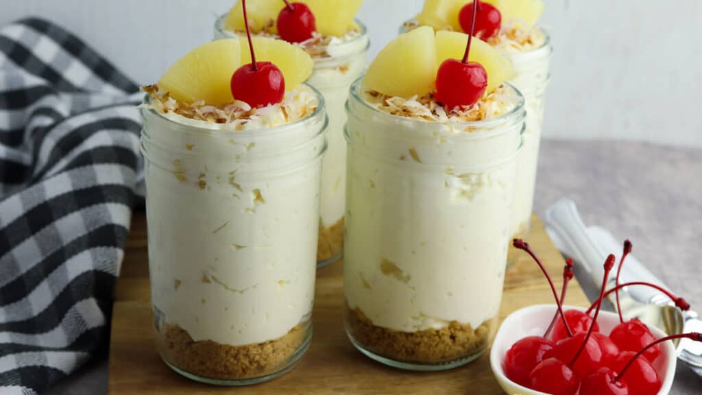 Four jars of pina colada cheesecake on a wooden cutting board topped with pineapple, coconut and cherries.