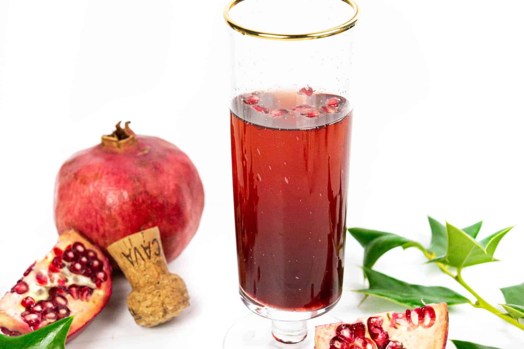 A pomegranate mimosa in a champagne flute next to holly, pomegranates and a champagne cork.