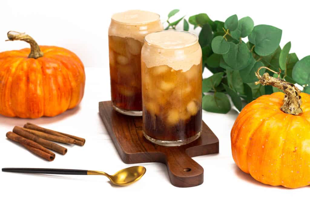 Two pumpkin cream cold brews sitting together on a dark brown wooden cutting board with decorative pumpkins to either side along with several cinnamon sticks, a black and gold spoon and some green leaves.