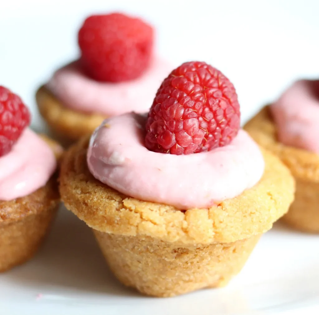 Closeup of cookie cups topped with icing and a raspberry on a white plate.