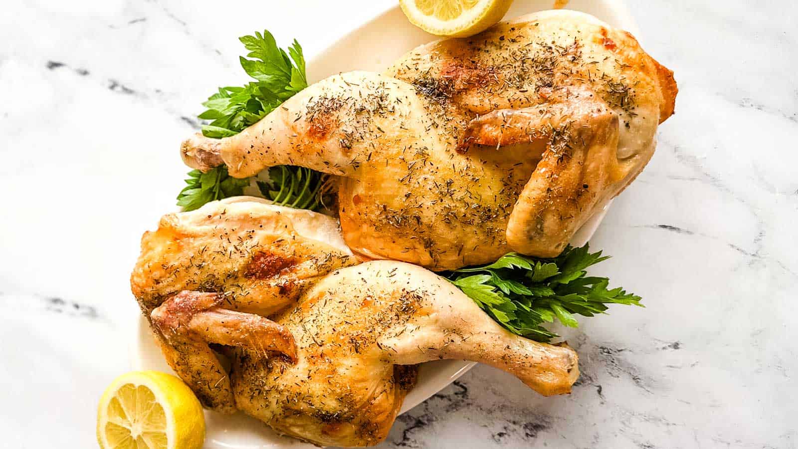 Two roasted chicken halves on a white platter with lemon and parsley.