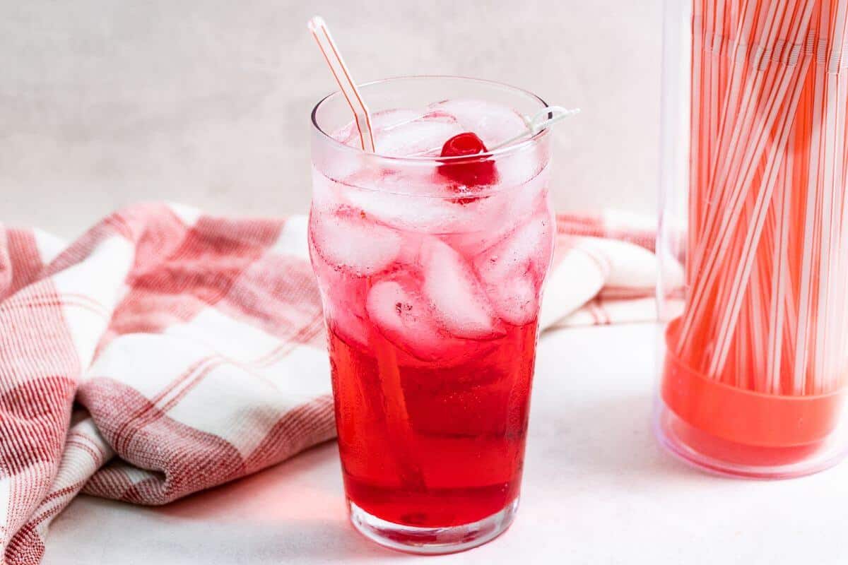 Shirley temple drink in tall glass with cherry garnish and straws container.