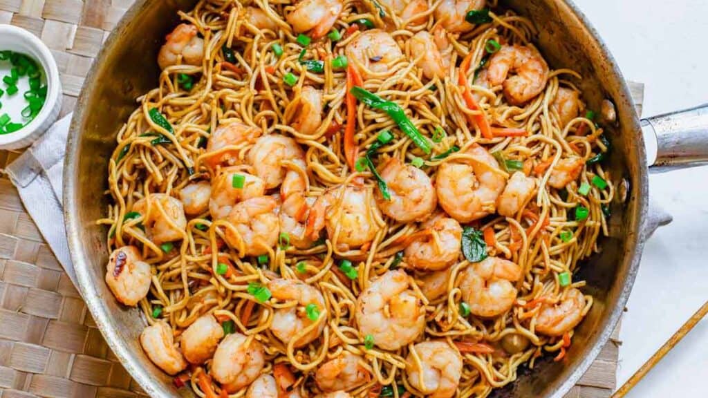 Shrimp lo mein in a skillet with chopped green onions in a white bowl on the side.