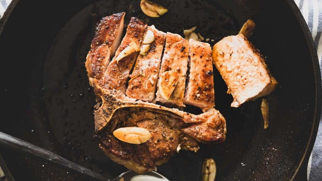 Bone in pork chop sliced in a skillet with garlic cloves.