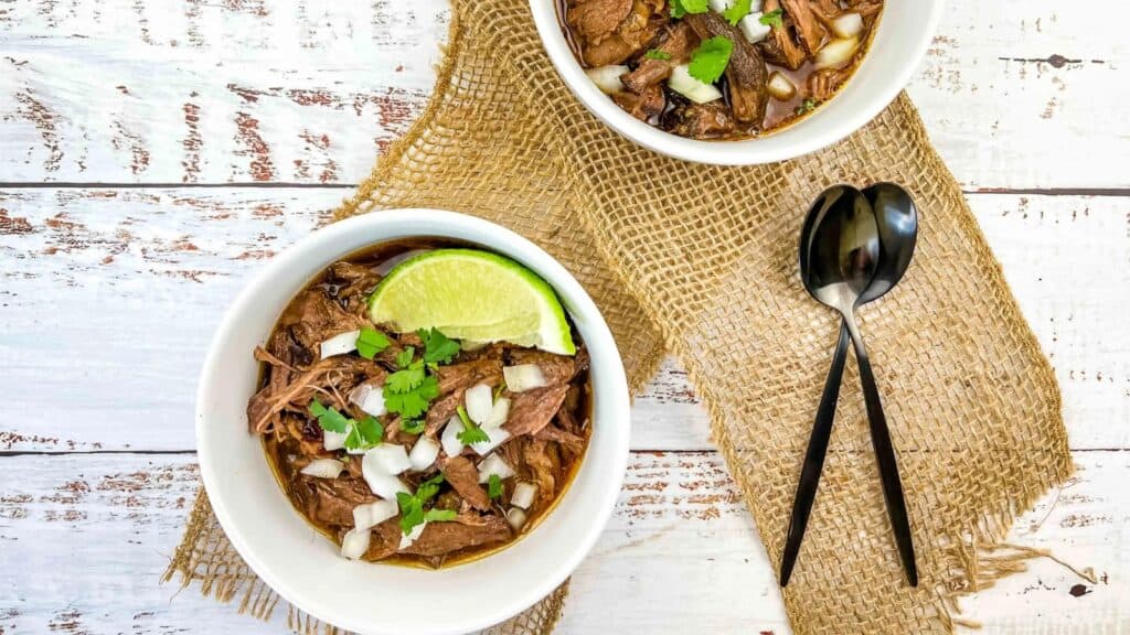 Two white bowls filled with slow cooker birria garnished with fresh lime wedges.