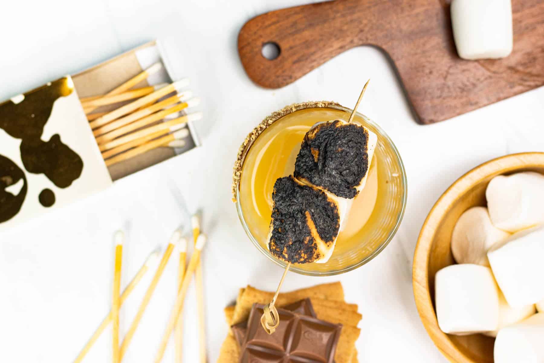 S'mores ingredients and matches surrounding a cocktail with charred marshmallows.