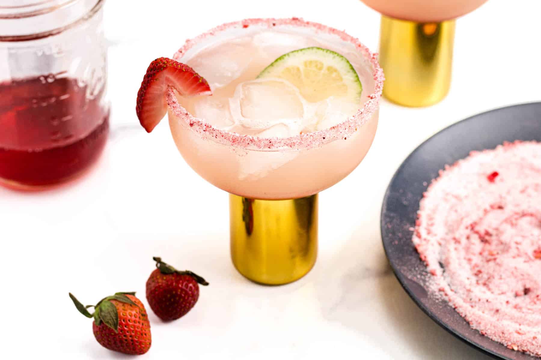The makings of a Strawberry Margarita sit on a white marble surface around the finished cocktail.