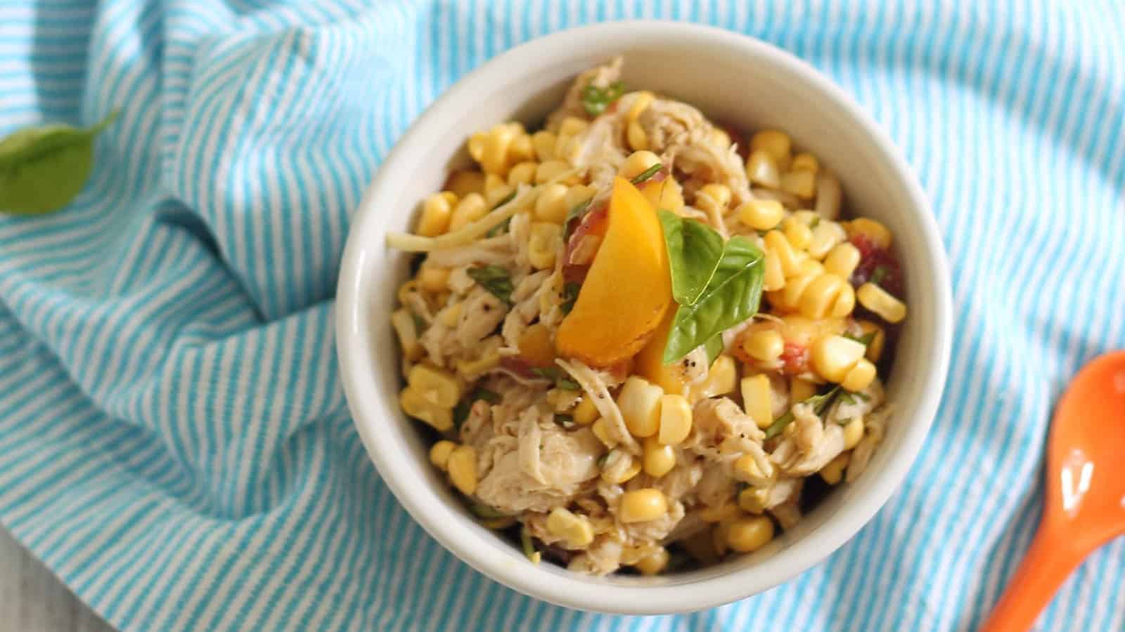 Sweet corn peach chicken salad with honey mustard dressing in a small white bowl with orange spoon and basil garnish.
