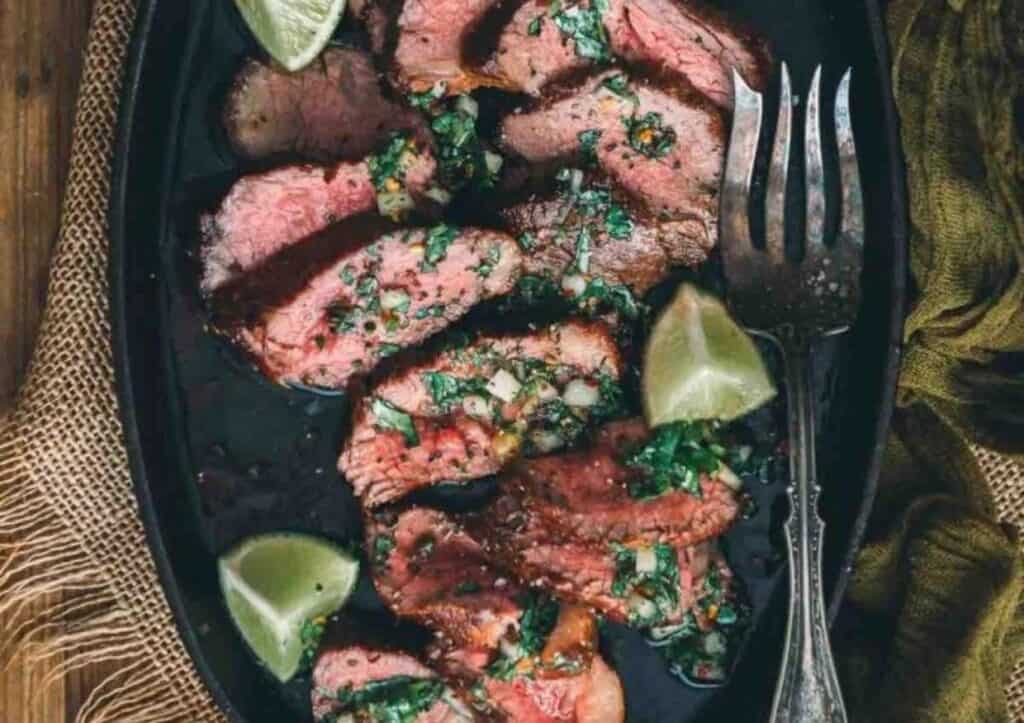 Sliced steaks on a board with sauce and lime wedges.