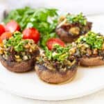 A white plate with vegan stuffed portobello mushrooms garnished with cherry tomatoes and parsley.