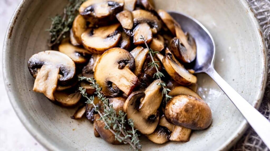 A tan ceramic bowl filled with sauteed mushrooms garnished with a fresh thyme sprig.