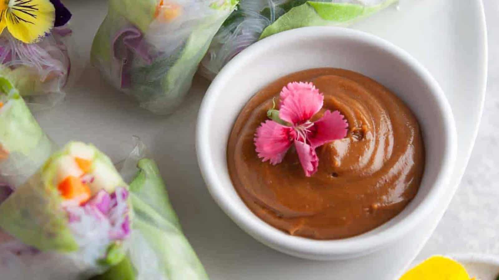 A bowl of Vietnamese peanut sauce with an edible flower and fresh rolls on the side.