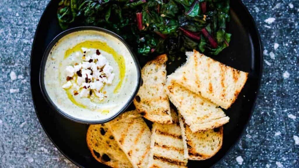 A bowl of feta cheese dip with bread and greens on a black plate.