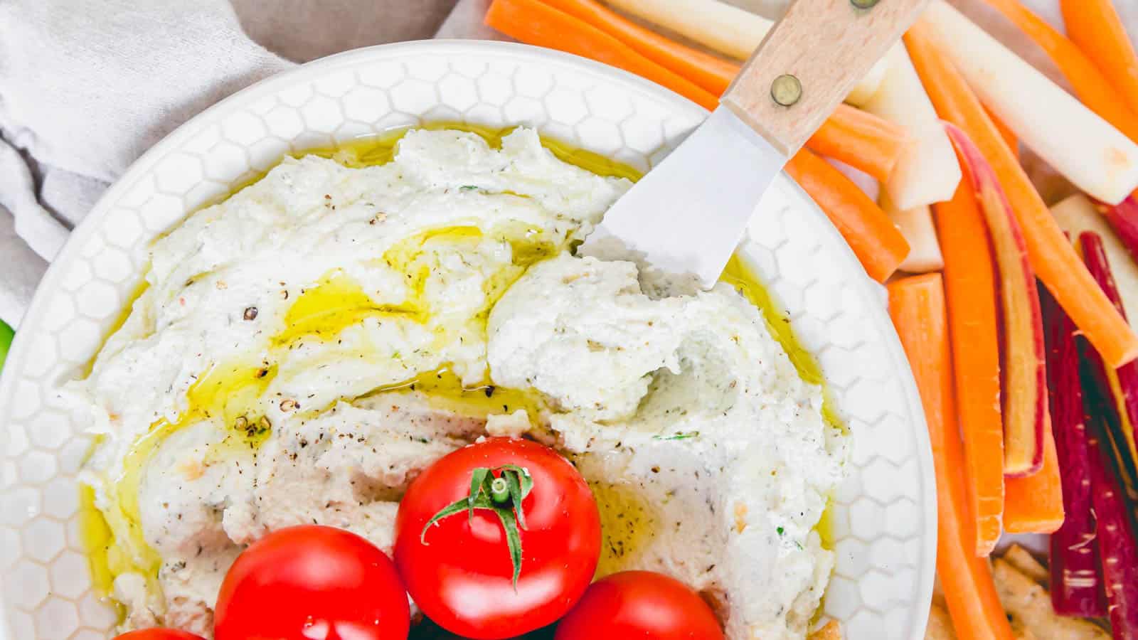 Whipped goat cheese dip topped with olive oil in a bowl with cherry tomatoes and a cheese spreader.
