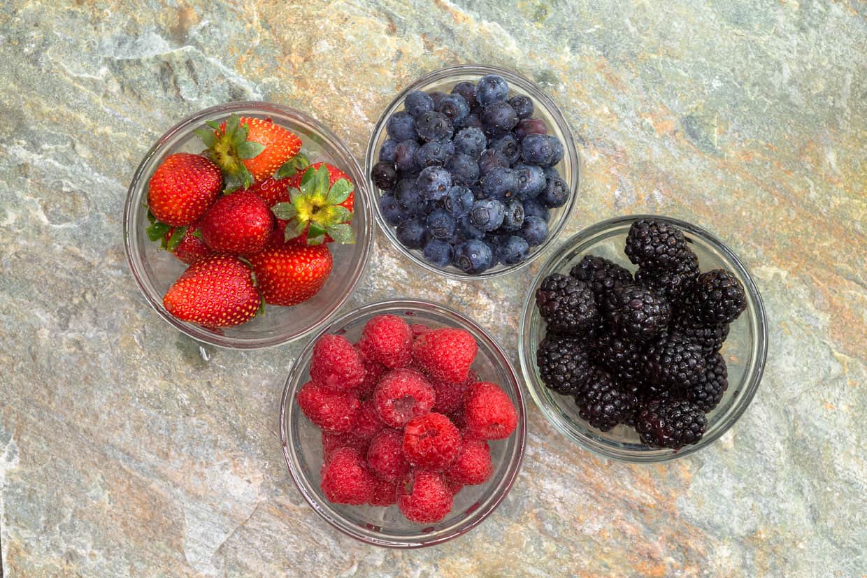 Four bowls of berries on a stone surface.