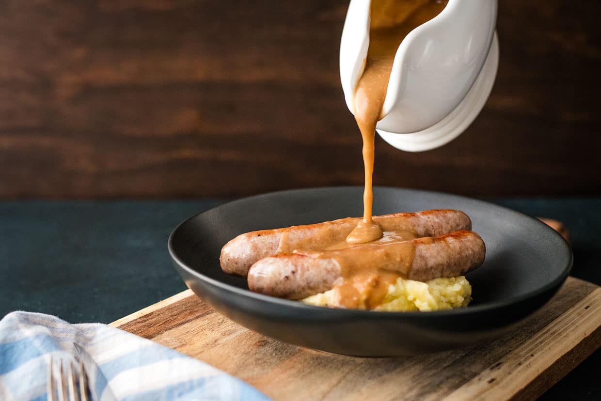 Gravy being poured over sausages and mashed potatoes.