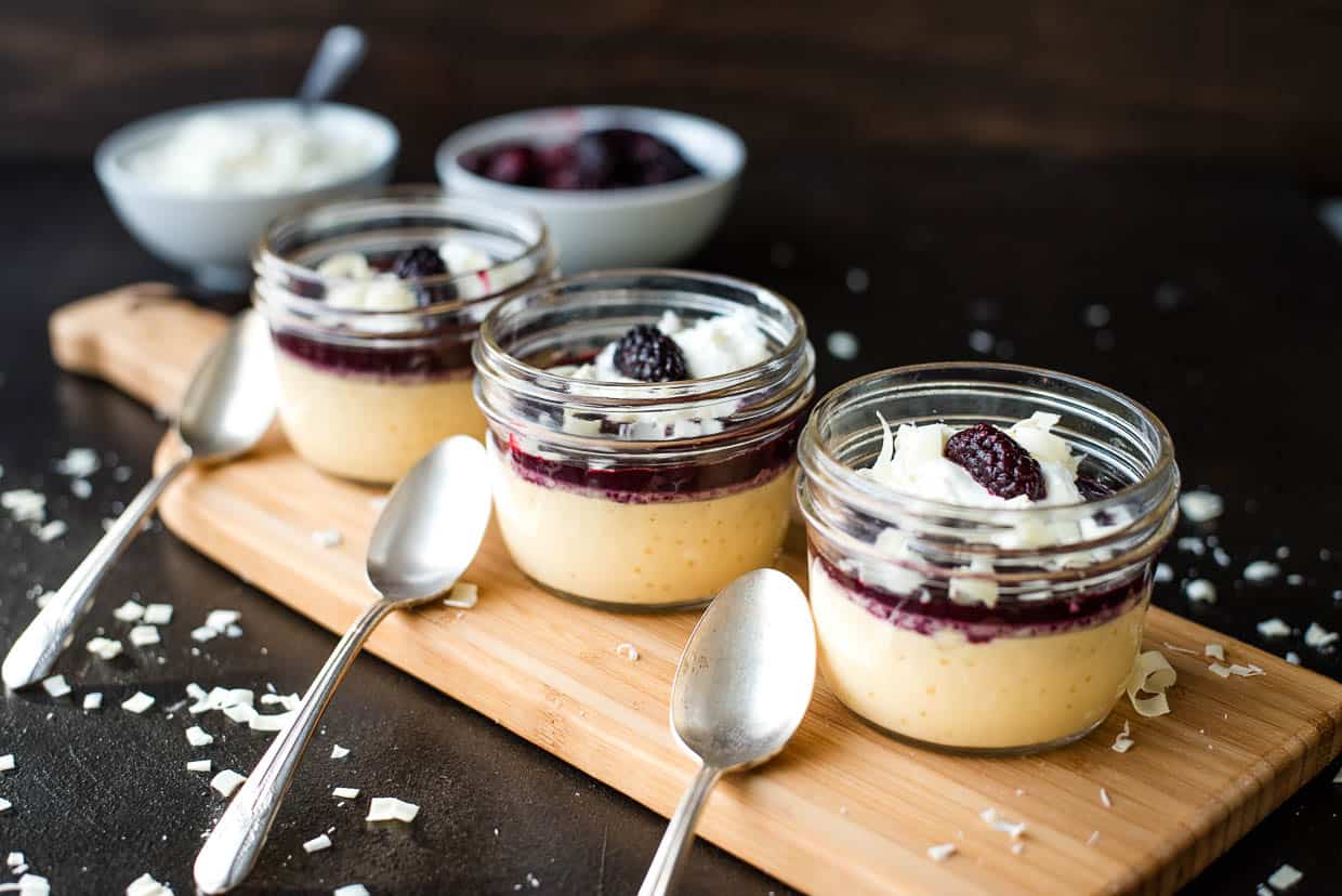 Three jars of custard dessert topped with a blackberry sauce with spoons on a wooden cutting board.