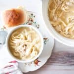 A bowl of chicken and noodle soup with noodles and a roll.